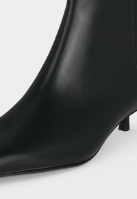 Bottine noire en cuir avec bout pointu et talon court et étroit sur fond blanc.