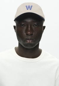 Light beige cap with a blue "W" logo, paired with a textured white long-sleeve shirt. Simple design, no additional accents or patterns.