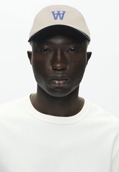 Casquette beige clair avec un logo bleu "W", associée à une chemise blanche à manches longues texturée. Design simple, sans accents ni motifs supplémentaires.