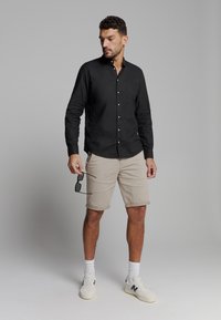 Zwarte longsleeve button-up shirt, kaki shorts, witte sneakers met marineblauwe accenten, houdt een zonnebril vast, effen achtergrond.