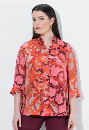 Frau mit langen dunklen Haaren, die Creolen trägt und eine mehrfarbige Blumenbluse in den Farbtönen Rot, Rosa und Orange trägt, steht vor einer hellen Wand.