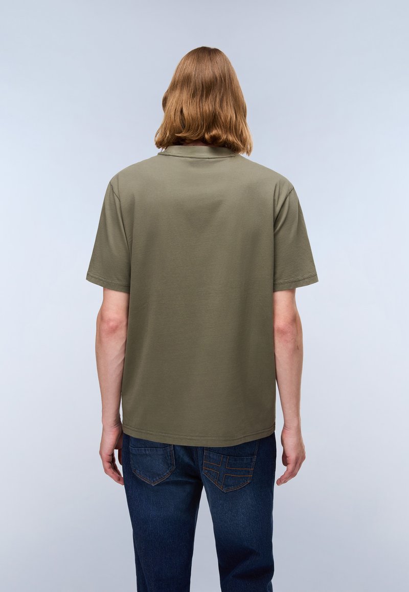 Oliwkowa bawełniana koszulka z luźnym krojem i krótkimi rękawami, przedstawiona z tyłu. Nosi ją w zestawieniu z ciemnoniebieskimi dżinsami.