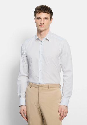 Junger Mann, der ein hell gestreiftes weißes Hemd und beige Hosen trägt, steht vor einem schlichten weißen Hintergrund.