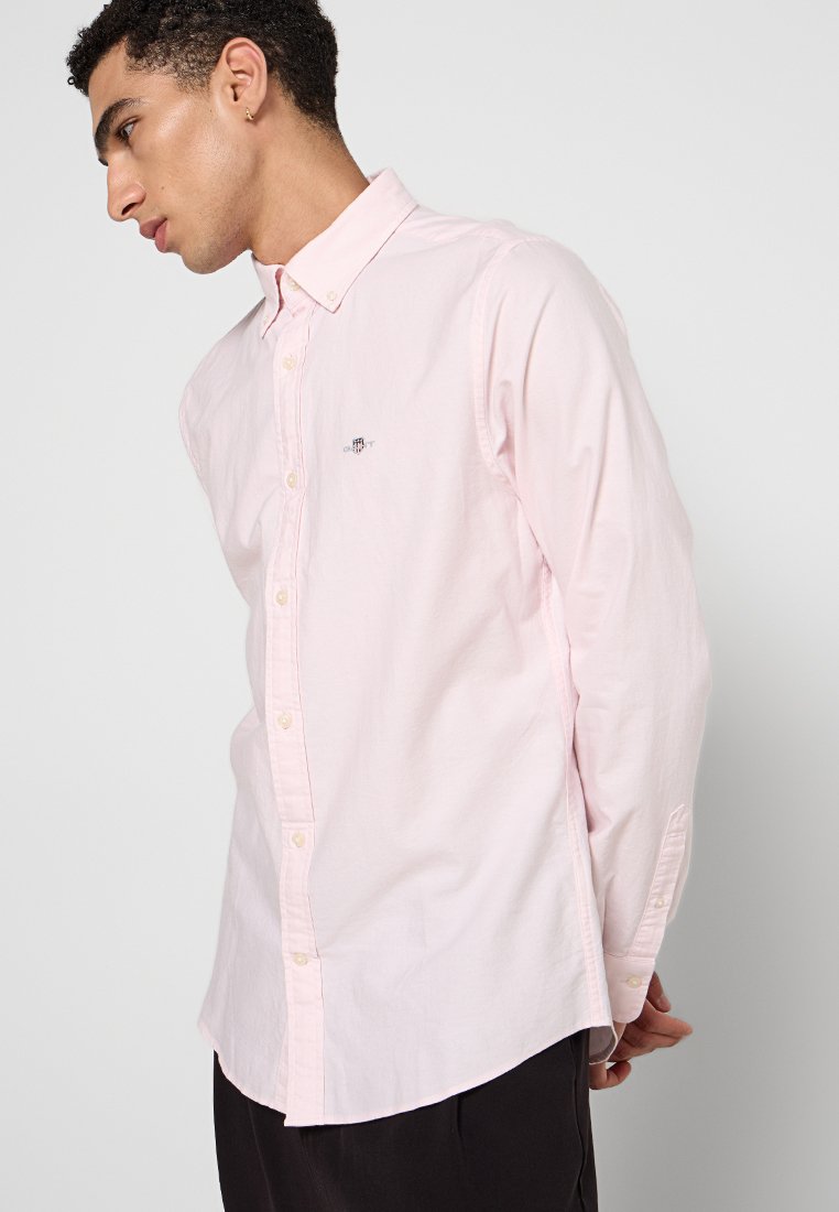 Mann mit kurzen lockigen Haaren, der ein hellrosa Hemd mit Knöpfen trägt und zur Seite blickt, mit hinter dem Rücken verschränkten Händen. Schlichter Hintergrund.