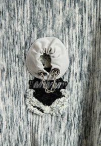 Tejido texturizado en tonos gris y negro con una sección circular fruncida que presenta un anillo de metal y una cadena, junto a un detalle bordado en contraste.