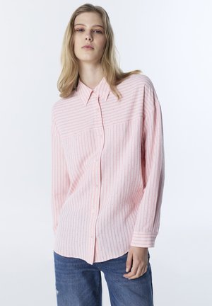Jeune femme portant une chemise ample rose clair à boutons avec des rayures verticales et des points, associée à un jean bleu, sur un fond blanc uni.