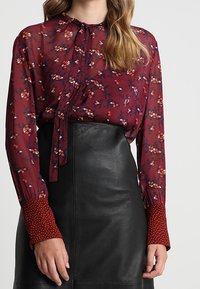 Blusa trasparente bordeaux con stampe floreali, dotata di un nodo al collo e polsini a pois, abbinata a una gonna di pelle nera.