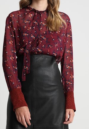 Blusa trasparente bordeaux con stampe floreali, dotata di un nodo al collo e polsini a pois, abbinata a una gonna di pelle nera.