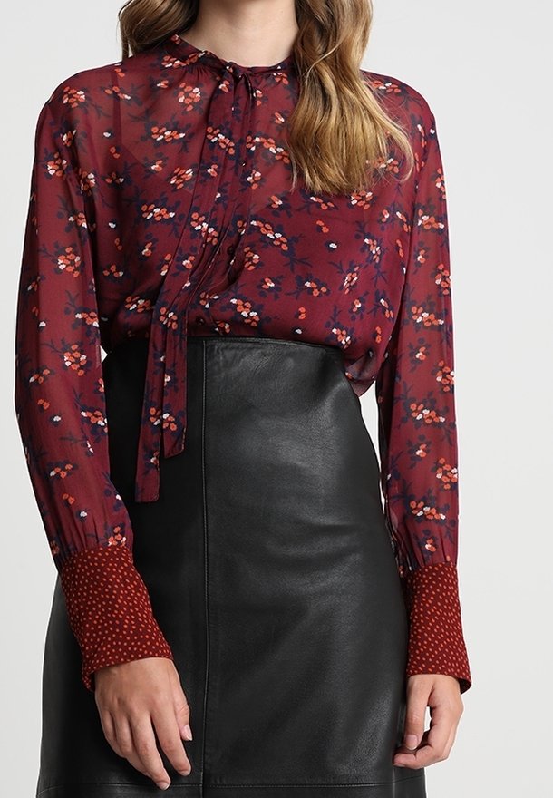 Blusa trasparente bordeaux con stampe floreali, dotata di un nodo al collo e polsini a pois, abbinata a una gonna di pelle nera.