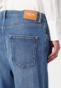 Siniset farkut korkealla vyötäröllä, joissa on ruskea nahkalette, johon on painettu "REPLAY", ja takataskussa hienovarainen punainen ommeltu yksityiskohta.