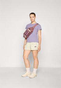 Sac banane Patagonia en bordeaux avec une sangle réglable, porté par-dessus une chemise lilas et un short beige clair. Des baskets et chaussettes beiges complètent le look.