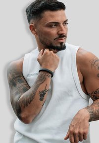 Man with trimmed beard and earrings, wearing white sleeveless shirt, showing detailed tattoos on both arms, looking to the side thoughtfully.