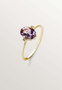 Anillo de oro con una gema ovalada púrpura, sostenida por cuatro garras, con una banda lisa y un acabado pulido.