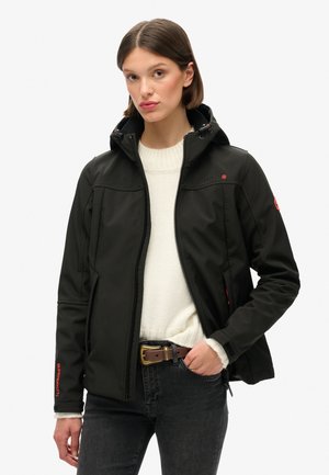 Black softshell jacket with a fitted design, zippered pockets, adjustable hood, and subtle red accents. Worn over a white sweater.