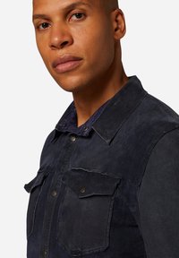 Mörk denimjacka med krage, knappstängning fram, två bröstfickor och en lätt sliten textur.