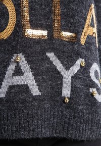 Grijze gebreide trui met glanzende gouden en zilveren paillettenletters, versierd met kleine gouden studs op de stof.