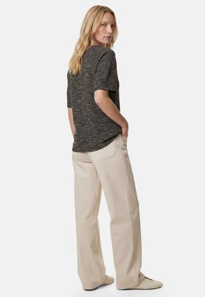 Frau mit blondem Haar trägt ein dunkles, strukturiertes Kurzarmoberteil und hellbeige weit geschnittene Hose, steht mit den Händen in den Taschen und schaut über die Schulter.