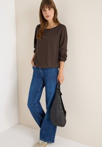 Blusa marrone a maniche lunghe con polsini elastici, abbinata a jeans svasati azzurri chiari. Una borsa a tracolla trapuntata nera è tenuta nella mano sinistra.