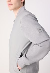 Jachetă bomber gri, cu un corp neted și mâneci tricotate texturate. Include buzunare laterale și o etichetă cu logo pe mâneca stângă.