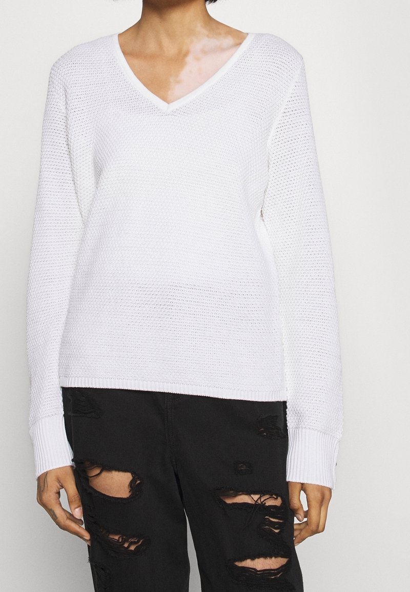 Pull blanc en maille avec col en V et manches longues, doté de poignets côtelés. Porté sur un jean noir déchiré avec de grandes déchirures.