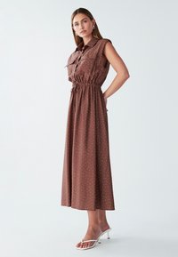 Brązowa, bezrękawnikowa sukienka maxi z kołnierzykiem i białymi kropkami. Posiada elastyczny pas i zwiewną spódnicę. Noszona z białymi sandałami.