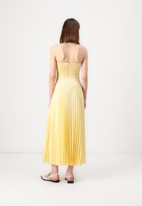 Fållat gult strapless-klänning med en figurnära överdel och svävande kjol. Bakre dragkedjestängning, tillsammans med ljusfärgade flata sandaler.
