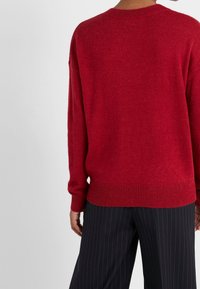 Pull en tricot rouge à coupe décontractée, col rond et poignets côtelés. Le tissu semble doux avec une texture subtile, associé à un pantalon à rayures.