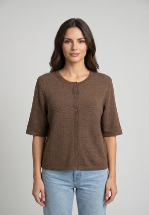 Donna con capelli scuri che indossa un cardigan marrone a maniche corte con bottoni e jeans azzurri su uno sfondo chiaro uniforme.