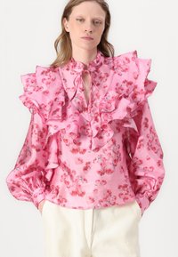 Blusa floreale rosa con maniche arricciate, scollatura a chiave e una texture satinata liscia. Il design presenta piccoli fiori rossi e accenti verdi.