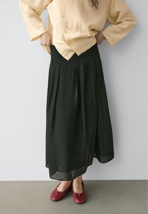 Personne portant une longue jupe noire plissée, une chemise jaune ample boutonnée, et des chaussures plates rouges, debout contre un mur uni.