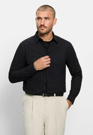 Mann mit kurzen Haaren, der ein schwarzes Hemd mit Knöpfen und beige Hose trägt, richtet sein Hemd mit einer Hand, während die andere in der Tasche ist.