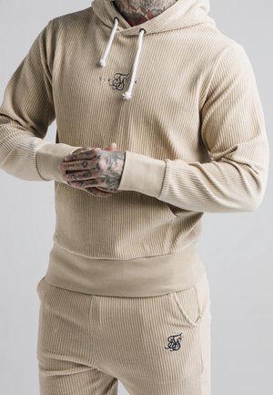 Person iført beige ribbet hættetrøje og matchende bukser med synlige tatoveringer på hænder og nakke, stående med hænderne foldet sammen.