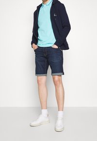 Sweat à zip bleu, polo bleu clair, shorts en denim foncé avec des revers, baskets blanches et chaussettes blanches. Tenue simple et décontractée.