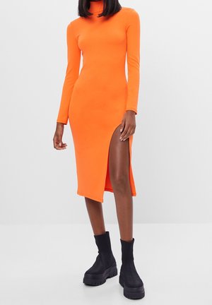 Frau trägt ein leuchtend orangefarbenes Midi-Kleid mit langen Ärmeln und seitlichem Schlitz, kombiniert mit schwarzen klobigen Plateau-Stiefeln vor weißem Hintergrund.