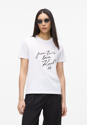 Femme aux longs cheveux foncés portant des lunettes de soleil noires, un t-shirt blanc avec le texte "de Paris avec amour, Karl" et un pantalon noir sur un fond uni.