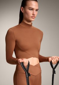 Bruine lange mouwen crop top met hoge hals en hoekige uitsnijdetails, gecombineerd met zwarte weerstandsbanden die een gladde textuur benadrukken.