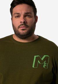 Olivově zelené bavlněné tričko s kulatým výstřihem; na levé straně hrudníku má opotřebovaný zelený grafický motiv "M+". Hladká textura, volný střih.