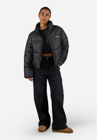 Svart dunjakke med høy krage og glidelås, båret over en svart crop top, kombinert med brede svarte jeans og brune støvler.