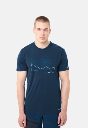 Mann trägt ein marineblaues T-Shirt mit weißem Berglinien-Design und Logo, steht vor einem schlichten hellgrauen Hintergrund.