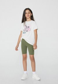 Biała koszulka z różowym graficznym nadrukiem aparatu, w połączeniu z oliwkowymi szortami rowerowymi i białymi trampkami. Ubranie ma luźny krój.