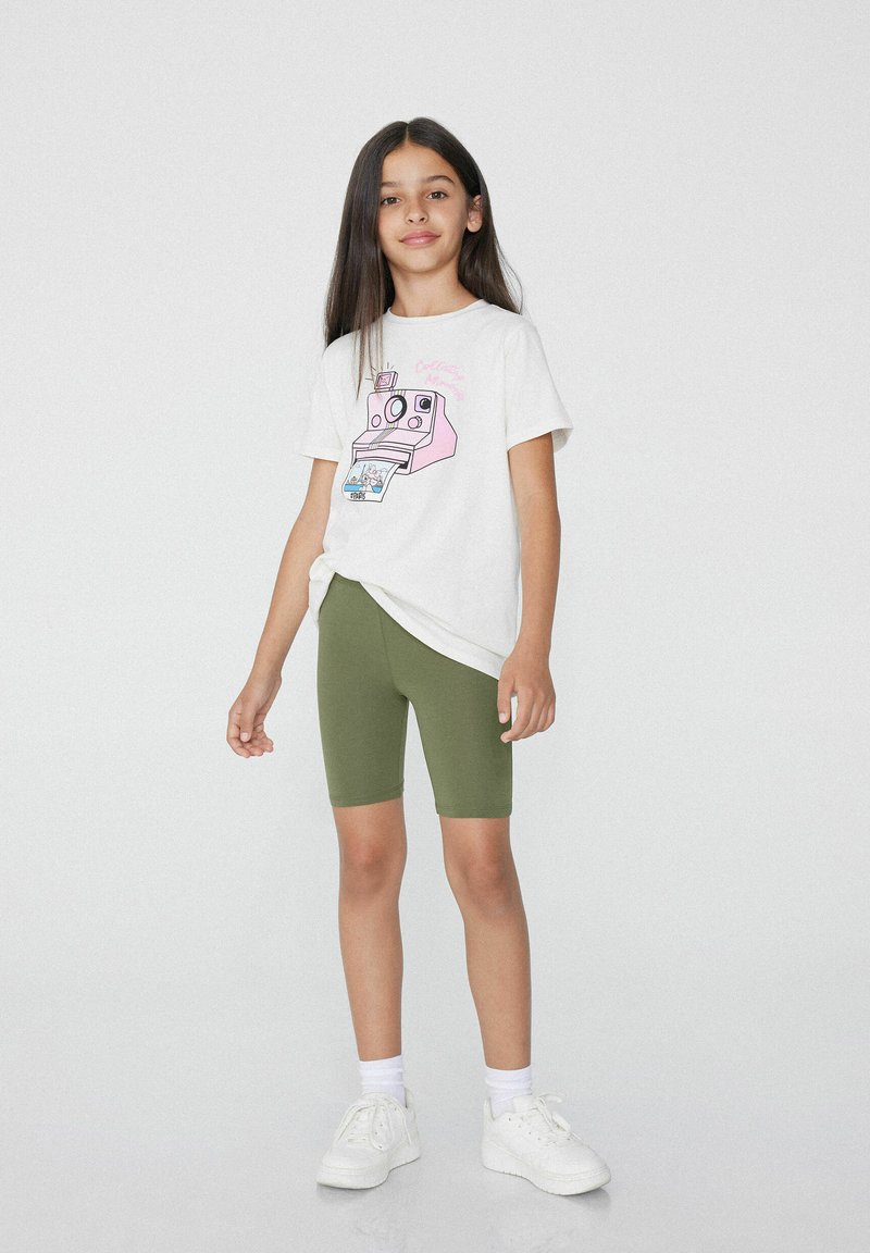 Biała koszulka z różowym graficznym nadrukiem aparatu, w połączeniu z oliwkowymi szortami rowerowymi i białymi trampkami. Ubranie ma luźny krój.