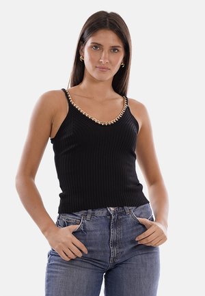 Donna con lunghi capelli castani indossa un top nero senza maniche a coste con dettaglio catena dorata e jeans blu a vita alta, mani in tasca.