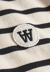 Sūnu audums krēmkrāsā un melnā krāsā ar apļa izšūtu pazīmi, kurā melnā diegā ir burts "W" uz teksturētas virsmas.
