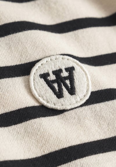 Tkanina w paski w kolorze kremowym i czarnym, z okrągłą haftowaną naszywką z literą "W" w czarnym nici na teksturowanej powierzchni.