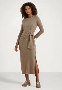 Moteris su garbanotais plaukais vilkinti taupe spalvos midi suknelę su ilgomis rankovėmis, šonu plyšta ir mazgu, derinant su taupe spalvos sportiniais bateliais.