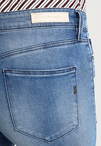 Siniset farkkutrikoot, joissa on takatasku ompeludetaljilla ja pieni logotunniste, viimeistelty haalistuneella pesulla.