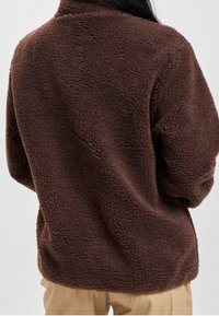 Giacca di pile marrone con superficie testurizzata, vestibilità comoda e colletto alto. Presenta spalle scese e senza chiusure visibili.