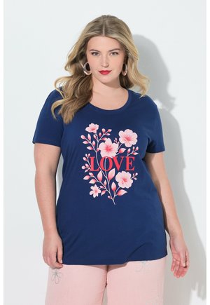 Kvinde med blondt hår iført øreringe i hoop-stil, marineblå T-shirt med pink blomsterdesign og teksten "LOVE", kombineret med lyserøde bukser.