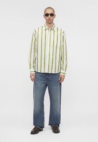 Homme portant des lunettes de soleil, une chemise blanche à rayures verticales vertes, un jean bleu ample et des chaussures marron foncé, debout devant un fond uni.