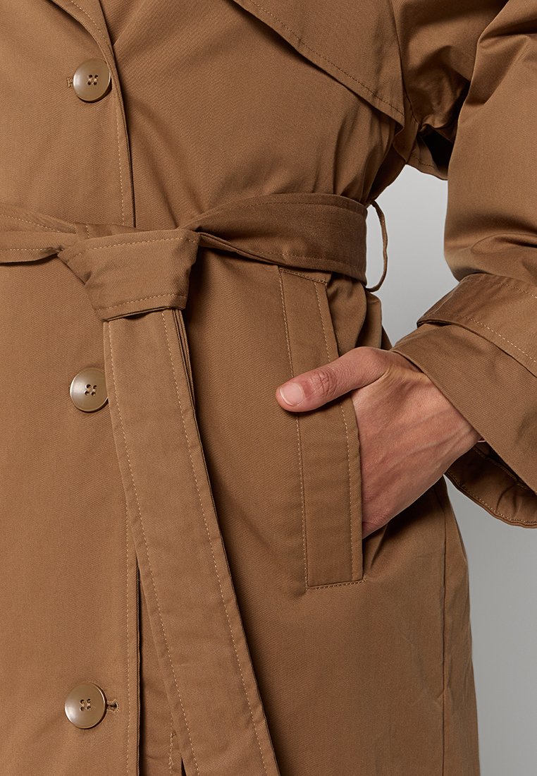 Manteau trench brun, texturé, avec une ceinture, des poches latérales et de grands boutons. Présente un large col et une coupe ajustée.